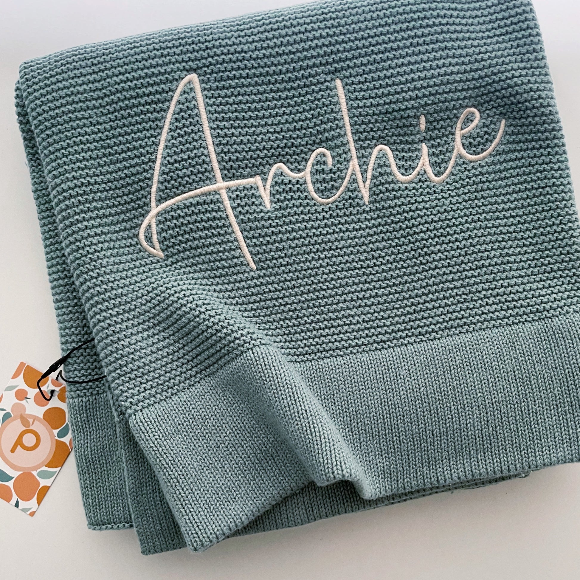 Seamist green coloured folded cotton blanket with the name Archie stitched onto it.