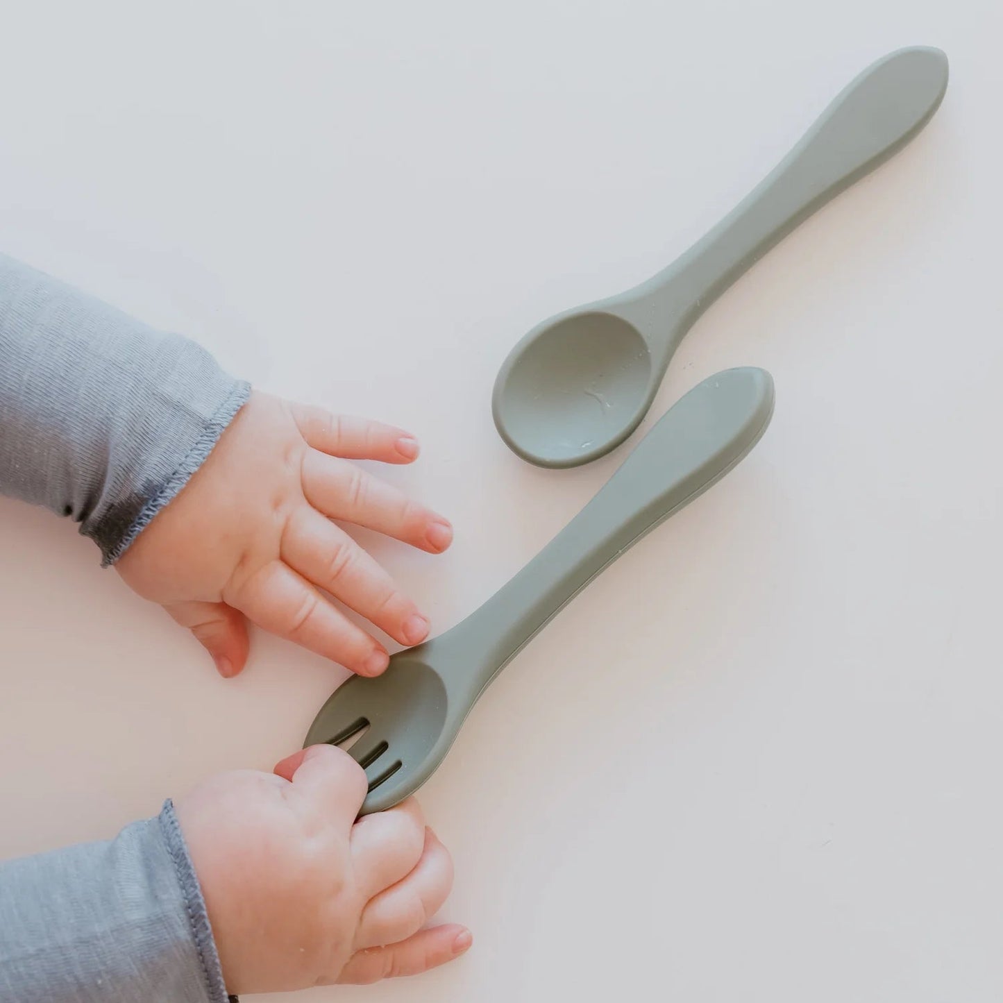 Two green plastic spoons on a light surface with a child's hand reaching towards them.