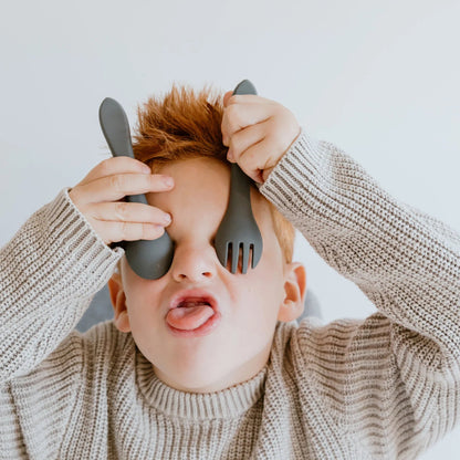 Child wearing a knit sweater holding kitchen utensils over their eyes against a plain background